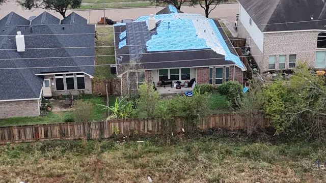 Drone view from behind a second tornado-damaged home in Cypress, where wind took off so much of the roof that entire slopes had to be dried in with blue synthetic underlayment instead of spot patches. The open field and damaged trees behind the fence line show how exposed these homes were to the storm. A full-slope dry-in protects the attic and ceilings until the roof can be completely replaced through the insurance claim.