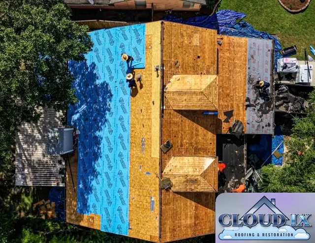 Straight-down drone view of a wind and hail claim reroof in Cypress, Texas. The old shingles and felt are stripped off, exposing the roof decking, with fresh OSB installed on both dormers and blue synthetic underlayment already going down on one side. Tarps protect the landscaping and the neighbor's side of the property from debris. Seeing the decking exposed like this is the only way to find and replace soft or damaged wood before the new roof goes on.
