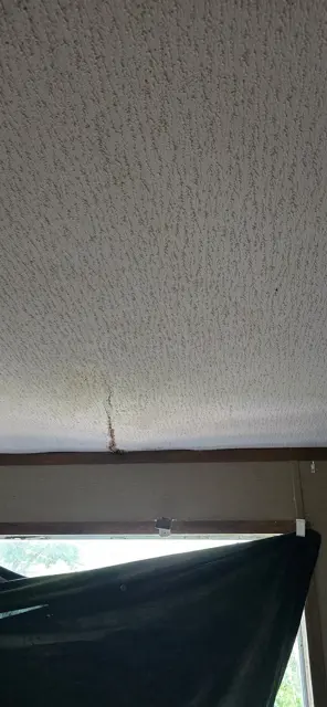 A crack runs through the textured ceiling with a brown water stain along it, marking where leaking rainwater collected above the drywall. Ceiling stains and cracks are often the first sign homeowners notice of a roof leak, sometimes weeks after the storm that caused it. Photographing them right away helps connect the interior damage to the roof damage in the insurance claim.