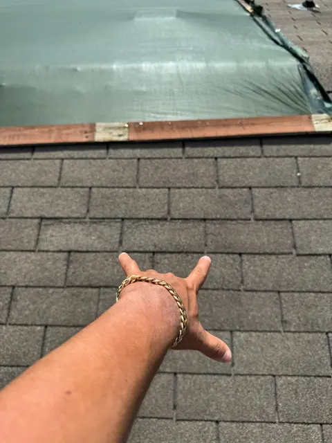 Wind-lifted shingles along the edge of the emergency-tarped section, photographed with a hand for scale. Neighboring tabs show the same lifting pattern, which indicated damage across the whole roof instead of a single blown-off spot. Evidence spread across multiple slopes is often what moves a claim from a small spot repair to a full roof replacement, so Cloud IX documents every slope during the inspection.