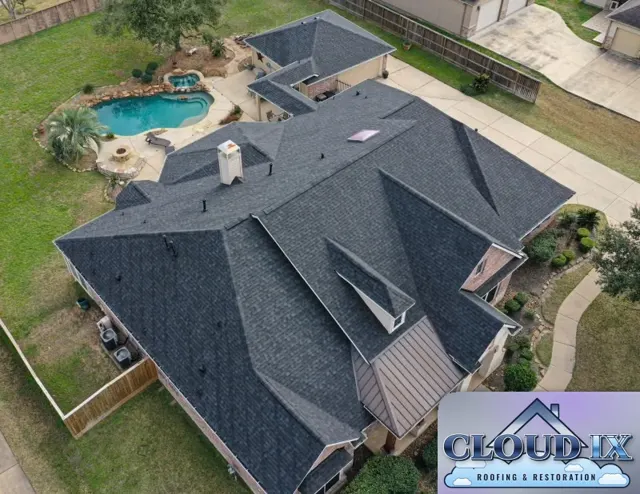 The finished Tomball hail claim reroof seen from the drone: Class 3 impact-rated architectural shingles in a dark charcoal color cover the complex hip roof, with crisp ridge caps along every hip and clean lines through the valleys. Class 3 shingles are rated under the UL 2218 impact test to withstand a 1.75-inch steel ball dropped from height, a step up in hail resistance over standard shingles for Houston-area homes. The chimney, skylight, roof vents and the bronze standing-seam metal roof over the front porch are all tied in.