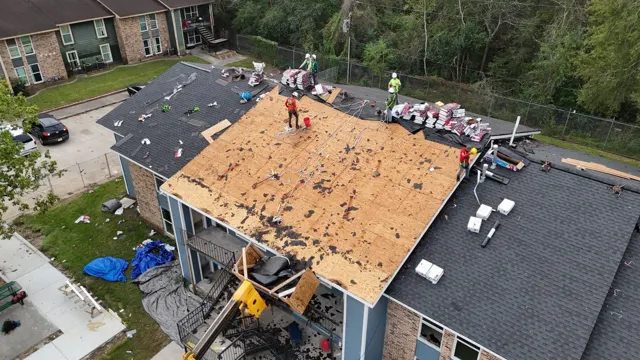 Close aerial of the roofing crew on a freshly torn-off section of the apartment roof. Every worker is tied off with a harness and rope to anchors at the ridge, and shingle bundles and underlayment rolls are staged along the peak, ready to go down. The neighboring roof sections already have new shingles, showing how the job moves building by building across the complex.
