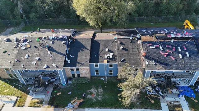 Overhead view of the connected roof sections on the two-story apartment building. The crew shingles one roof while the next is dried in with dark synthetic underlayment and stocked with bundles. Long multi-family buildings have many roof-to-roof transitions, and each one needs proper flashing and shingle tie-ins so the finished roof works as one watertight system.