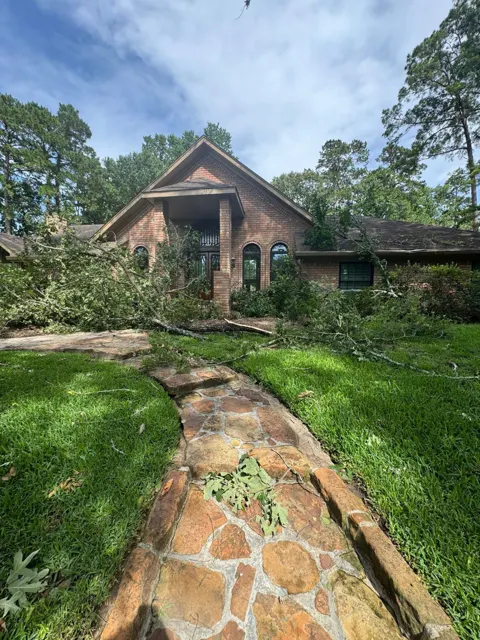 Wind storm damage at a brick home in Tomball, Texas: large oak limbs snapped off in high winds and came down across the roof, the front entry and the landscaping, blocking the stone walkway to the front door. Tall pines and hardwoods surround the property, which is common in Tomball and why tree damage is one of the most frequent storm calls in the area. Before any roof repair can start, the limbs have to be safely cleared so the full roof can be inspected.